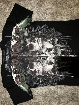 Svart DeVille t-shirt med döskalletryck - Unik svart t-shirt från DeVille med stort grafiskt tryck av döskalle, änglar och gröna detaljer på framsidan. T-shirten har korta ärmar och coolt tryckt logga i nacken. Materialet känns som mjuk bomull och passformen är normal.