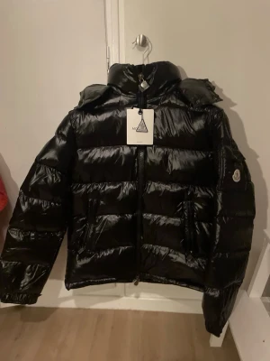 Svart Moncler pufferjacka med huva - Säljer en svart, glansig pufferjacka från Moncler med huva och dragkedja framtill. Jackan har två fickor med dragkedja och en Moncler-logga på ärmen. Perfekt för kalla dagar och riktigt snygg till streetwear-stilen. Motsvarar en storlek medium 