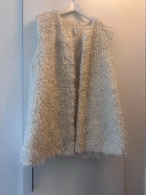 Vit fluffig pälsväst från L.O.G.G. - Säljer en supermjuk och fluffig vit pälsväst från L.O.G.G. Västen har en öppen front och är gjord i ett lurvigt material som ger en riktigt cozy vibe. Perfekt att slänga över en hoodie eller tröja för extra värme och stil.