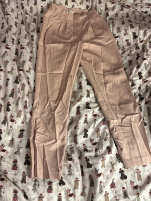 Beiga kostymbyxor från Vila - Säljer ett par snygga beiga kostymbyxor från Vila i storlek 36. Byxorna har hög midja, raka ben och pressveck framtill. Tillverkade i ett mjukt och lätt material som känns skönt mot huden. Perfekta till en stilren look.