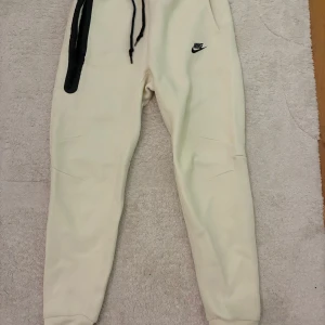 Nike Tech mjukisbyxor - beiga, nya - Säljer ett par beige Nike TECH mjukisbyxor med svart logga på vänster lår och svarta snören i midjan. Byxorna har en relaxed passform och är tillverkade i mjukt material som känns skönt mot huden. Endast testade. Storlek S