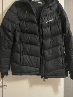 Svart pufferjacka från Everest, strl 38 - Säljer en svart pufferjacka från Everest i storlek 38. Jackan har huva med snörning, dragkedja framtill och flera praktiska fickor, både på utsidan och insidan. Perfekt för kalla dagar och riktigt skön att bära. Klassisk och stilren design med Everest-logga på bröstet. den har en liten defekt och det är ett litet snitt vid armen
