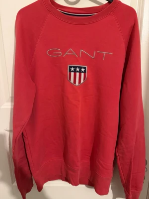  GANT tröja  - Röd sweatshirt från GANT i bomull med broderad logga och sköld med stjärnor och ränder på bröstet. Klassisk rund halsringning och långa ärmar. Perfekt för dig som gillar sportig och stilren look.