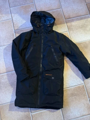 Svart parkas från Berkeley Gore-Tex - Säljer en svart parkas från Berkeley med Gore-Tex-material. Jackan har huva, två stora fickor med lock och knappar, samt två snedställda bröstfickor med dragkedja. Insidan är fodrad i grått och har detaljer i orange. Perfekt för kalla och regniga dagar. Använd 1 gång! Nyskick. Nypriset 4990:- 