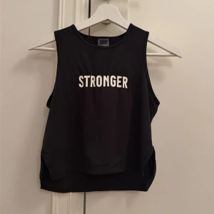 Svart linnetopp från Stronger - Säljer en svart linnetopp från Stronger med vit text framtill. Toppen är ärmlös och har en loose passform, perfekt för träning eller sportiga aktiviteter. Materialet är mjukt och lätt polyester som andas bra.