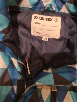 Blå vinteröverall Snowtex mönstrade - Säljer en vinteröverall från Snowtex med coolt geometriskt mönster i blå, svart och vit. Byxorna har dragkedjor, resår vid benslut och är gjorda i slitstarkt syntetmaterial. Perfekt för vinteraktiviteter och håller dig varm och torr.
