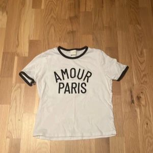 Vit t-shirt med svart text från H&M - Vit t-shirt i storlek S från H&M med svart text 'AMOUR PARIS' på bröstet. Svarta kontrastdetaljer vid halsringning och ärmslut ger en cool vibe. Klassisk rund hals och korta ärmar. Perfekt för dig som gillar enkel men snygg stil. Använt flertal gånger men bra skick, säljer den för att jag har växt ur den. Har en jätte ljus liten fläck vid texten men behövde kolla nära för att se den. Jag kan absolut skicka bild på fläcken ifall det behövs.