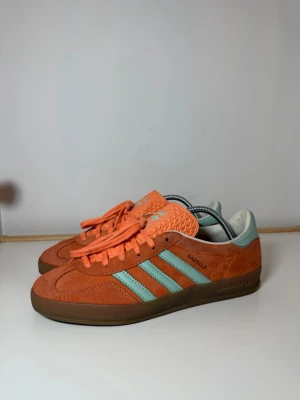Adidas Gazelle orange och turkos - Säljer ett par Adidas Gazelle sneakers i orange mocka med turkosa detaljer och klassiska tre ränder på sidan. Skorna har snörning, platt sula och rund tå. Insidan är vit och yttersulan är brun. Perfekt för dig som gillar färgstarka och unika sneakers.