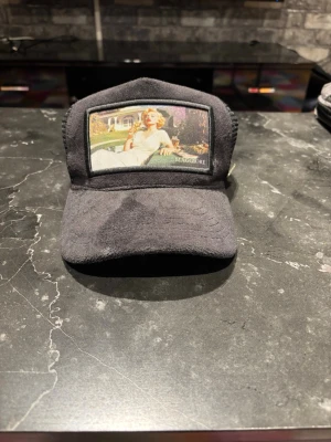 Svart truckerkeps från Maggiore - Säljer en svart truckerkeps från Maggiore med mesh på sidorna och baksidan. Framtill finns en patch med ett konstmotiv och logotypen. Kepsen har justerbar snapback och en liten metallogo på sidan. Perfekt för att lyfta din streetwear-look.