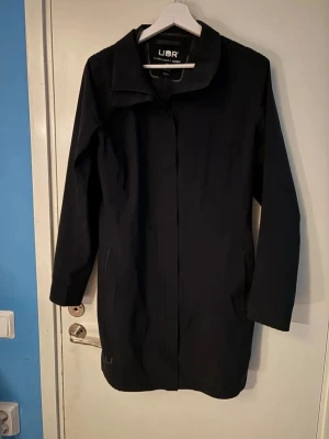 Svart rock från UBR, storlek M - Säljer en stilren svart rock från UBR i storlek medium. Jackan har en klassisk krage, diskret logga vid nederkanten och dragkedja framtill. Materialet är syntet och designen är enkel och clean, perfekt för dig som gillar minimalistisk stil.