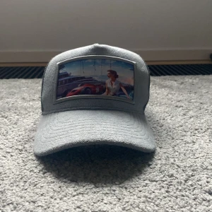  Ljusblå revenite keps  - Säljer en grå truckerkeps med mesh på sidorna och baksidan. Framsidan har ett färgglatt bildmotiv med bil och kvinna. Kepsen har böjd skärm och justerbar snapback. Perfekt för dig som vill sticka ut med en unik accessoar.