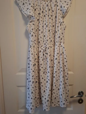 Vit blommig klänning från H&M - Superfin vit klänning från H&M med svart blommigt mönster. Klänningen har korta volangärmar, rund hals och snörning i midjan för en snygg passform. Materialet är lätt och luftigt, perfekt för varmare dagar. Passar dig som gillar en feminin och avslappnad stil.