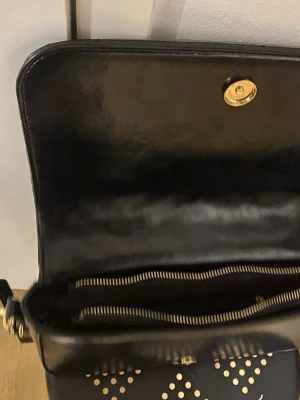 Svart quiltad axelväska från V 1969 - Snygg svart axelväska i glansigt skinn med quiltad yta i sicksackmönster och guldfärgade detaljer. Väskan har ett stort V 1969-tryck på framsidan, justerbar axelrem och dragkedja inuti. Perfekt för dig som gillar stilrena accessoarer med lite edge.