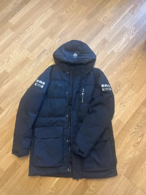 Svart vinterjacka från Sail Racing strlk M - Säljer en svart vinterjacka från Sail Racing med huva och coola loggor på ärmarna. Jackan har stora fickor framtill, dragkedja och tryckknappar. Snyggt Sail Racing-tryck på bröstet och detaljerad badge.