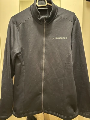 Mörkblå fleecejacka från J.Lindeberg - Mörkblå fleecejacka från J.Lindeberg med hel dragkedja och hög krage. Diskret logga på bröstet och ett stort grafiskt tryck på ryggen. Jackan har långa ärmar och är perfekt för kyliga dagar. Materialet är mjukt och bekvämt.