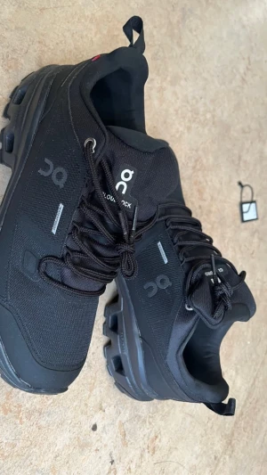 Svarta On Cloudrock sneakers - Säljer ett par svarta On Cloudrock sneakers med grov sula och snörning. Skorna är tillverkade i slitstark syntet och har vattentätt material. Logga på sidan och plös, samt detaljer i grått. Perfekta för dig som gillar outdoor och vill ha stilrena skor med robust känsla.