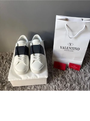 Valentino Garavani vita sneakers - Säljer ett par snygga vita Valentino Garavani sneakers med svart detalj över snörningen. Skorna har rund tå, platt sula och är tillverkade i skinn. Kommer med originalpåse och certifikat. Perfekt för dig som gillar exklusiva märken och stilren design.