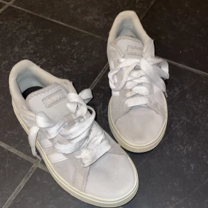 Adidas sneakers i ljusgrå mocka - Säljer ett par Adidas sneakers i ljusgrå mocka med vita ränder på sidorna och klassisk gummisula i beige. Skorna har rund tå, platt sula och snörning. Perfekt för dig som gillar stilrena och tidlösa sneakers med retrovibe. Säljer för att dem är för små för mig.
