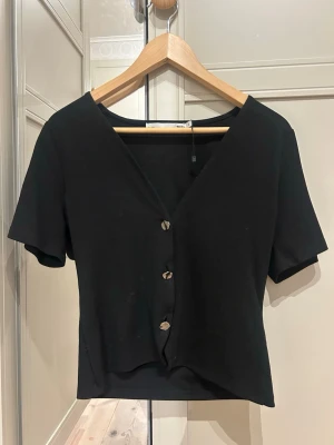 Svart kortärmad blus med knappar - Säljer en svart blus med korta ärmar och v-ringning. Toppen har fyra dekorativa knappar framtill och är lätt croppad i modellen. Perfekt till jeans eller kjol för en enkel men snygg look.