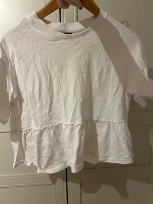 Vit volangblus från Gina Tricot - Säljer en vit blus från Gina Tricot med rund hals och korta ärmar. Toppen har en volangdetalj nedtill som ger en söt och avslappnad look. Materialet är mjuk bomull och modellen är normal i passningen. Perfekt för sommarens alla tillfällen!