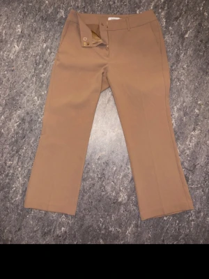 Beiga bootcut kostymbyxor - Snygga beiga kostymbyxor med bootcut-snitt och klassisk midja. Byxorna har knapp och dragkedja framtill samt bälteshällor. Perfekta för dig som gillar stilrena och tidlösa plagg. Materialet känns mjukt och har en lätt stretch.