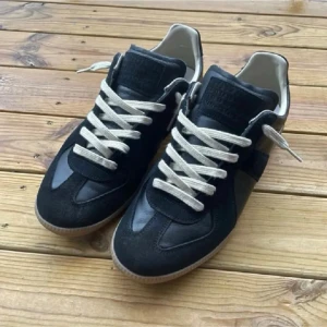 Svarta Maison Margiela sneakers - Maison Margiela sneakers i svart med beige snörning och gummisula. Skorna har en mix av mocka och skinn, rund tå och platt sula. Klassisk design med diskret logga på plösen och en grå detalj på hälen. Perfekta för dig som gillar stilrena och coola sneakers.