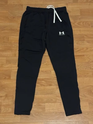 Svarta mjukisbyxor från Under Armour - Svarta mjukisbyxor från Under Armour med vit logga på vänster lår och vit snörning i midjan. Byxorna har en slim passform och är gjorda i mjukt bomullsmaterial, perfekta för chill eller träning. Elastisk midja och sidofickor för extra bekvämlighet. Har två styckna en storlek S och en Storlek M