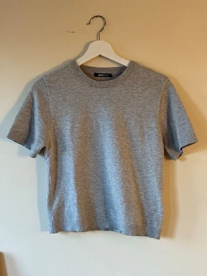 Grå stickad t-shirt från Gina Tricot - En enkel och stilren grå stickad t-shirt från Gina Tricot. Modellen har rund hals, korta ärmar och ribbade kanter vid hals, ärmslut och nederkant. Den har använts endast en gång och har inga skador. Perfekt för dig som gillar en clean look och mjuka material.