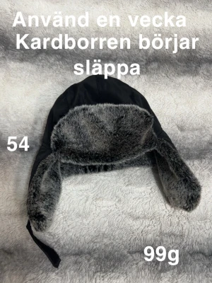 Svart pälsmössa med grått foder - Mysig svart mössa med grått fuskpälsfoder och öronlappar. Perfekt för kalla vinterdagar. Kardborreband för att fästa under hakan. Snygg och enkel design som håller dig varm.🌸Använd en vecka men resåren börjar släppa. Storlek 54  