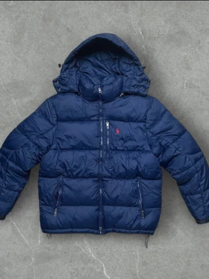 Blå pufferjacka från Polo Ralph Lauren - Säljer en mörkblå pufferjacka från Polo Ralph Lauren med huva och flera dragkedjor. Jackan har det klassiska röda Polo-logot på bröstet, snörning i huvan och quiltad design. Perfekt för kalla dagar och har en riktigt chill vibe.