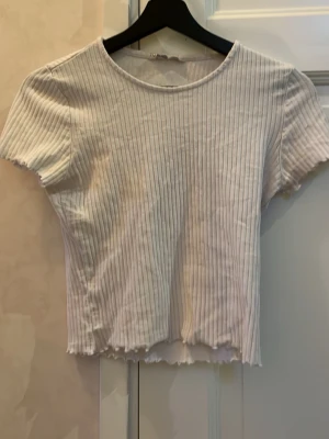 Vit ribbad topp från ONLY - Säljer en vit ribbad topp från ONLY i storlek S. Toppen har korta ärmar, rund halsringning och är croppad med vågig kant nedtill. Perfekt för dig som gillar enkel och stilren design med lite edge.