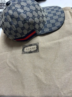 Svart Gucci keps med GG-mönster - Snygg svart keps från Gucci med klassiskt GG-monogram över hela kepsen. Har en grön och röd rand på sidan som ger den typiska Gucci-looken, kommer med tag o dustpåse