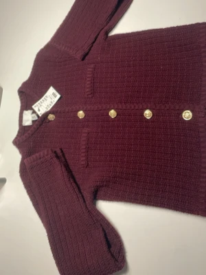 Vinröd stickad kofta med guldfärgade knappar - Säljer en vinröd stickad kofta från H&M med snygga guldfärgade knappar framtill. Koftan har två dekorativa fickor och långärmade ribbade ärmar. Perfekt för dig som gillar klassisk stil med en modern twist.