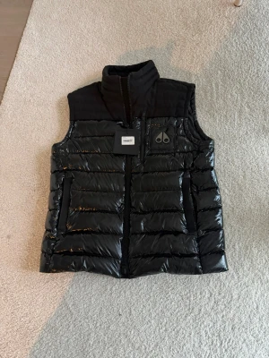 Moose Knuckles väst - Säljer en svart dunväst från Moose Knuckles, modell Dugald Vest. Västen har quiltad design med glansig yta, hög krage och dragkedja framtill. Märkets logga syns på bröstet. Perfekt för lager-på-lager under kyliga dagar.