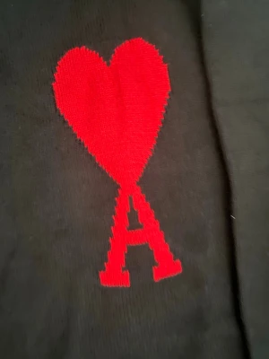 Svart stickad tröja från Ami Paris - Svart stickad tröja från Ami Paris med ikoniskt rött hjärta och bokstaven A broderat på bröstet. Klassisk rund halsringning och ribbade muddar vid ärmslut och nederkant. Perfekt för dig som vill ha en stilren men ändå statement look.