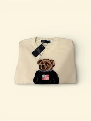 Vit stickad tröja Polo Bear Ralph Lauren - Vit stickad tröja från Polo Ralph Lauren med klassisk Polo Bear-motiv framtill. Tröjan har rund hals, ribbade muddar och lång ärm. Björnen bär en svart tröja med amerikansk flagga och jeans, samt RL-broderi vid nederkanten. Perfekt för dig som gillar ikoniska detaljer.