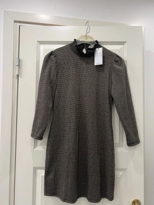 Rutig klänning Mango - Snygg kortklänning från Mango i svart och beige rutigt mönster. Den är helt oanvänd. Klänningen har långärmade ärmar, en volangdetalj vid kragen och en liten knappöppning bak i nacken. Materialet är mjukt. Perfekt för dig som gillar klassisk stil med en twist.