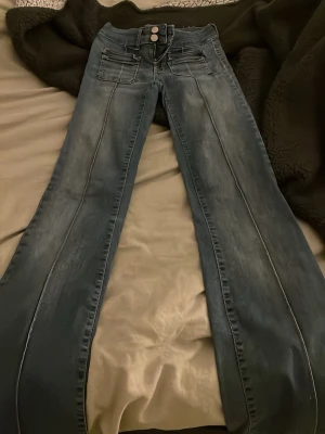 Blå bootcut jeans med dubbla knappar - Snygga blå jeans med bootcut passform och dubbla knappar i midjan. Jeansen har klassiska fickor fram och bak, samt slitna detaljer vid bensluten för en cool vibe. Materialet är jeans och färgen är en tvättad blå ton.