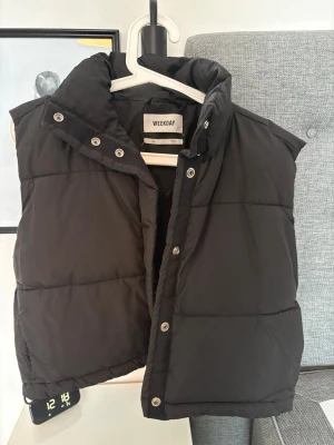 Svart dunväst från Weekday XS - Säljer en svart dunväst från Weekday i storlek XS. Västen har hög krage, tryckknappar framtill och quiltad design. Perfekt att slänga över en hoodie eller tröja när det är kyligt ute. Materialet är polyester och västen har en loose passform. Namn: cut puffer west 