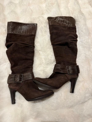 Bruna höga boots med flätning - Snygga mörkbruna boots i mocka med högt skaft och flätade detaljer upptill och runt ankeln. Skorna har en markerad klack och ett stort spänne. Dragkedja på insidan skon.