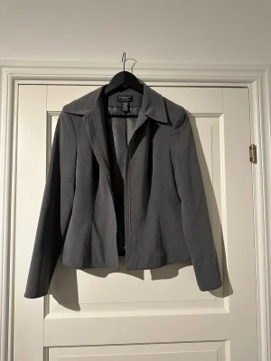 Grå kavajjacka från Worthington - Grå kavajjacka från Worthington med klassisk krage och diskret slits vid ärmslutet. Jackan har dragkedja framtill och är tillverkad i en mjuk stretchig polyesterblandning. Snygg, stilren och passar perfekt till en cool och dressad look.