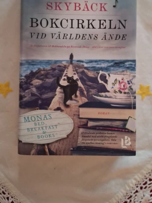 Bokcirkeln vid världens ände, bok - En charmig roman om vänskap, böcker och nya möten vid havet. Perfekt för dig som gillar mysiga berättelser, bokcirklar och oväntade äventyr. Omslaget lockar med en katt, te och böcker – en riktig feelgood för unga vuxna som vill drömma sig bort.