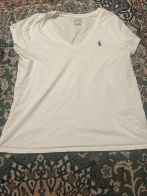 Vit v-ringad t-shirt från Polo Ralph Lauren - Vit t-shirt från Polo Ralph Lauren med v-ringning och klassisk blå logga broderad på bröstet. T-shirten är kortärmad och gjord i mjuk bomull, perfekt för en clean och enkel stil.