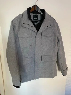 Grå ulljacka från Jack & Jones L - Snygg grå jacka från Jack & Jones i storlek L. Jackan har fyra fickor framtill, hög krage och quiltat svart innerfoder. Tillverkad i ullblandning med klassisk rak passform och diskreta knappar och dragkedja framtill.