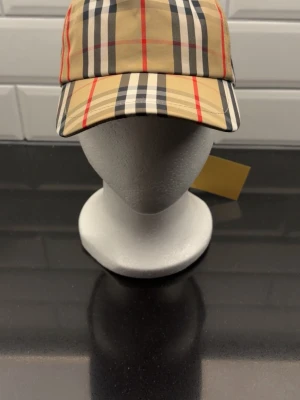 Rutig Burberry keps i beige och svart - Snygg keps från Burberry med klassiskt rutigt mönster i beige, svart, vitt och röda detaljer. Justerbar baktill med svart spänne och Burberry London England-märke. Perfekt accessoar för dig som gillar ikonisk design och streetstyle.