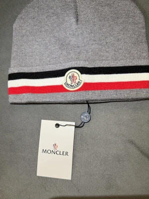Grå Moncler mössa med ränder - Snygg grå mössa från Moncler med klassiska svarta, vita och röda ränder samt Moncler-logga framtill. Mössan är ribbstickad och har en mjuk känsla, perfekt för att hålla värmen. Passar dig som vill ha en stilren accessoar med lyxig touch.