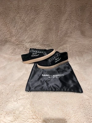 Svarta Saint Laurent sneakers - Säljer ett par svarta Saint Laurent sneakers med vit logga på sidan och beige platt sula. Skorna har snörning och en stilren design med rund tå. Materialet är en mix av skinn och textil, och medföljer gör en svart dustbag med Saint Laurent Paris-tryck.