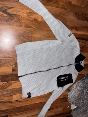 Grå kofta från Sail Racing med svart detalj - Riktigt snygg grå kofta från Sail Racing med svart krage och svart bröstficka med dragkedja. Ribbstickad design och hel dragkedja framtill.