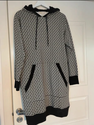 Svartvit hoodieklänning Michael Kors - Snygg svartvit hoodieklänning från Michael Kors med MK-logga över hela tyget. Klänningen har långa ärmar, huva med snörning och två stora fickor framtill med svarta detaljer. Mjuk och bekväm, perfekt för en chill och trendig look.