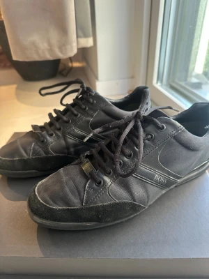 Svarta sneakers från Hugo Boss - Snygga svarta sneakers från Hugo Boss med stilren design. Skorna har snörning, rund tå och platt sula. Materialmix av skinn och syntet med Boss-logga på sidan och under sulan. Perfekta för dig som gillar klassisk streetstil.